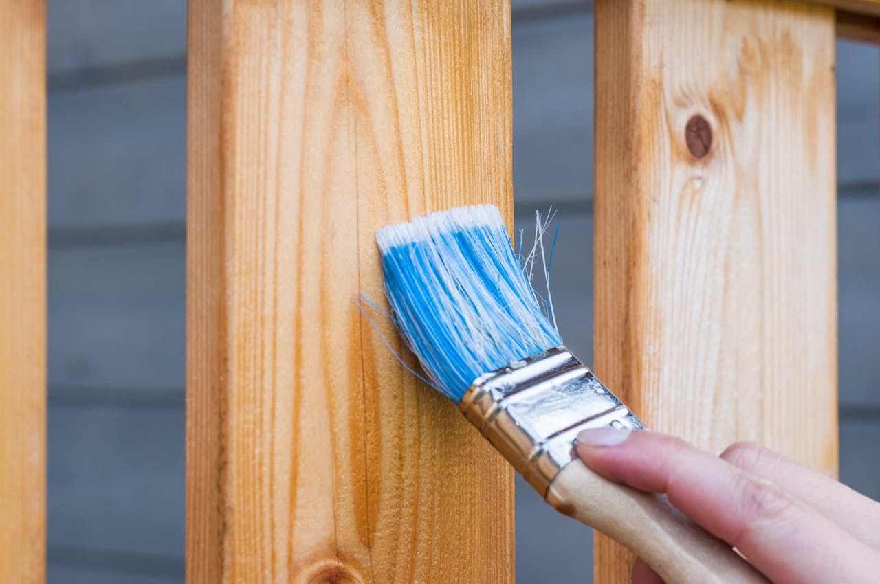 A hand painting a wooden fence plank with a blue paintbrush outdoors. Perfect for DIY and renovation themes.